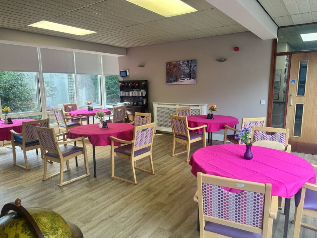 Dining room with round tables, covered brightly coloured table cloths, each with 3 chairs.